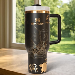 May include: Black Stanley travel mug with a gold-colored lid and handle. The mug features gold illustrations of characters and scenes from a movie, including a crab with the text "I'M SO SHINY". The Stanley logo is also in gold.