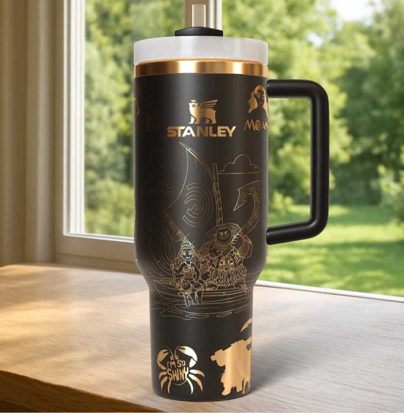 May include: Black Stanley travel mug with a gold-colored lid and handle. The mug features gold illustrations of characters and scenes from a movie, including a crab with the text "I'M SO SHINY". The Stanley logo is also in gold.