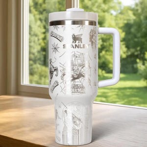 Puede incluir: Taza de viaje Stanley blanca con gr&aacute;ficos de Spider-Man. La taza tiene un asa y tapa blancas, con un borde plateado. El dise&ntilde;o incluye a Spider-Man, telara&ntilde;as y paisajes urbanos. La taza mide aproximadamente 23 cm de alto.