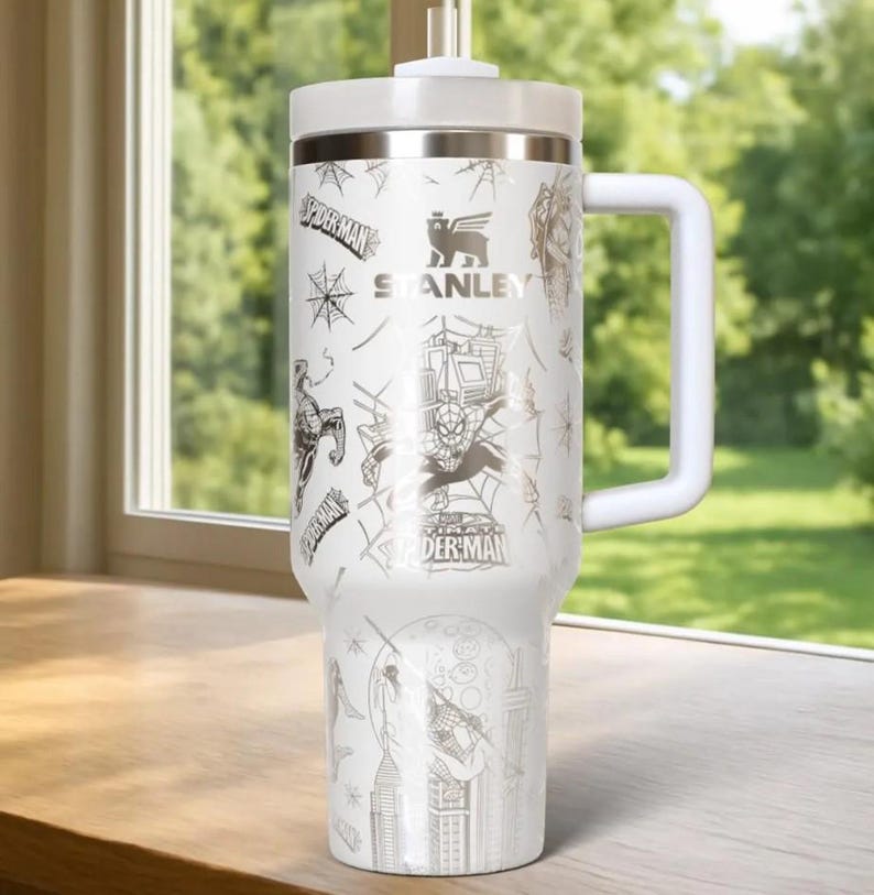 Puede incluir: Taza de viaje Stanley blanca con gr&aacute;ficos de Spider-Man. La taza tiene un asa y tapa blancas, con un borde plateado. El dise&ntilde;o incluye a Spider-Man, telara&ntilde;as y paisajes urbanos. La taza mide aproximadamente 23 cm de alto.