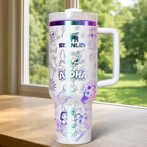 May include: White Stanley travel mug with a handle, decorated with a holographic design of Stitch and the word "Aloha." The mug has a white lid and a purple and green iridescent band. The Stanley logo is also visible.