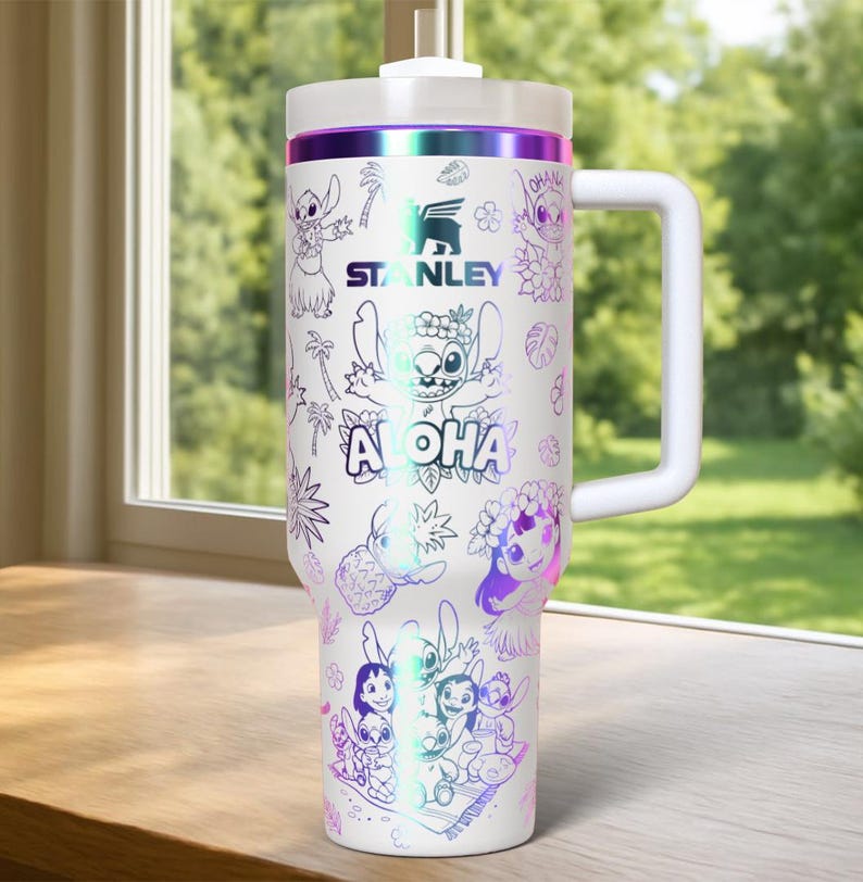 May include: White Stanley travel mug with a handle, decorated with a holographic design of Stitch and the word "Aloha." The mug has a white lid and a purple and green iridescent band. The Stanley logo is also visible.