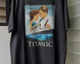 May include: Black t-shirt featuring a movie poster graphic from the film "Titanic." The graphic shows a couple embracing on the deck of the ship, with the word "TITANIC" in white block letters below the image. The shirt is hanging on a wooden hanger.