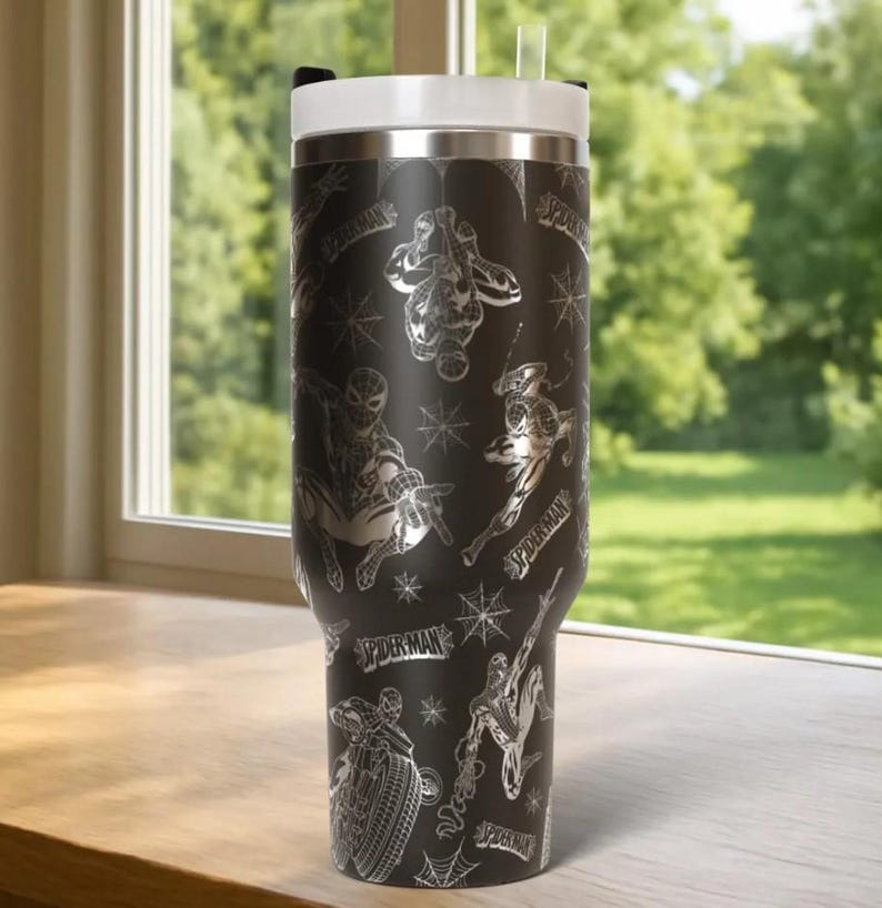 Puede incluir: Vaso t&eacute;rmico negro con ilustraciones plateadas de Spider-Man y telara&ntilde;as. Tiene una tapa blanca y una pajita transparente. Las palabras "Spider-Man" tambi&eacute;n son visibles en el vaso.