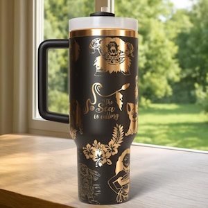 May include: Black travel mug with gold-colored illustrations of characters and floral designs. The mug has a black handle, a white lid, and the text "The Sea is Calling".
