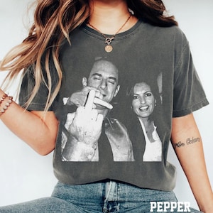 May include: Dark grey t-shirt featuring a black and white image of two people. One person is giving the middle finger. The text "PEPPER Color" is at the bottom of the shirt.