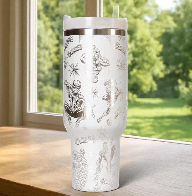 Puede incluir: Vaso t&eacute;rmico blanco con ilustraciones y logotipos de Spider-Man. El vaso tiene una tapa blanca y una banda plateada. Una pajita transparente est&aacute; insertada en la tapa. El dise&ntilde;o incluye im&aacute;genes de Spider-Man en varias poses.