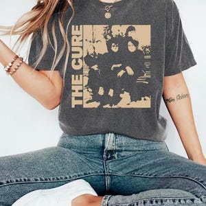 May include: Dark gray t-shirt featuring a beige graphic of a band and the text "THE CURE" in a vertical font. The shirt is made of a soft material and has a vintage look. The person in the image is wearing blue jeans and white sneakers.