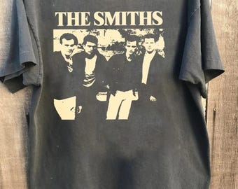 May include: Dark gray t-shirt featuring the band name "THE SMITHS" in white block letters above a black and white image of the band members. The shirt is on a wooden hanger.