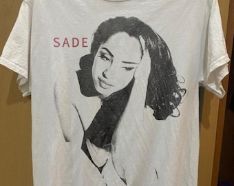 May include: White t-shirt featuring a black and white portrait of a woman with the name "SADE" printed in red above the image. The graphic tee has short sleeves and a crew neck.