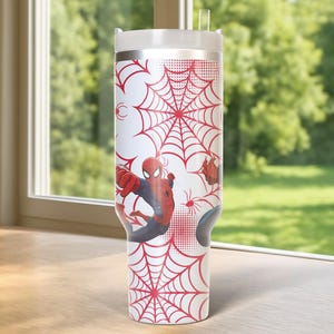 May include: A white tumbler featuring a red spiderweb design and illustrations of Spider-Man. The tumbler has a white lid and a clear straw. The design wraps around the entire tumbler, creating a cohesive look.