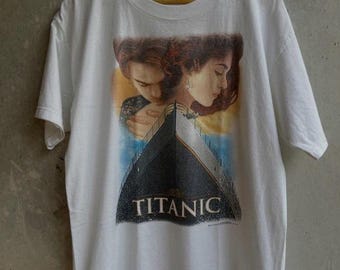 May include: White t-shirt featuring a graphic from the movie Titanic. The design includes a depiction of the ship and the faces of the main characters. The word "TITANIC" is printed in white on the ship's bow. The shirt is hanging on a wooden hanger.