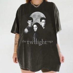 May include: Dark gray t-shirt featuring a graphic of three people in front of a full moon, with the text "the twilight saga" in white. The shirt has a vintage, worn look.