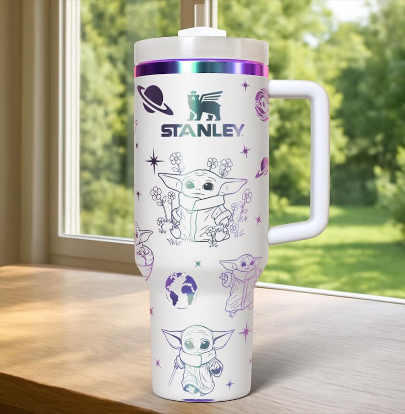 May include: White Stanley tumbler with a handle, featuring illustrations of a character and space-themed graphics. The tumbler has a white lid and a purple and green iridescent band. The Stanley logo is also visible.