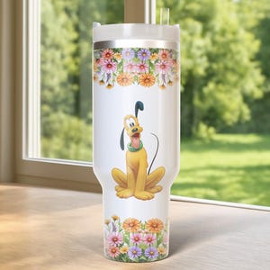 May include: A white tumbler featuring a cartoon image of Pluto, the Disney dog, with a floral design. The tumbler has a silver lid and a clear straw. The floral design includes pink, orange, and purple flowers. The tumbler is sitting on a wooden surface.