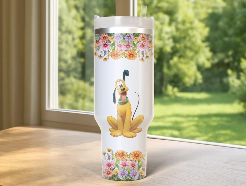 May include: A white tumbler featuring a cartoon image of Pluto, the Disney dog, with a floral design. The tumbler has a silver lid and a clear straw. The floral design includes pink, orange, and purple flowers. The tumbler is sitting on a wooden surface.