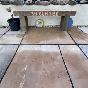 May include: A rectangular wooden bench with a light brown finish, featuring the text 34 ELMRISE carved into the front of the seat. The bench is supported by two thick, sturdy wooden legs and is positioned on a stone patio surface.