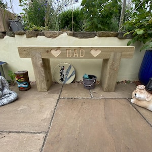May include: A light brown wooden garden bench featuring a horizontal seat supported by two thick legs. The front of the seat is engraved with the word DAD centred between two carved heart shapes. The bench has a natural wood grain finish.