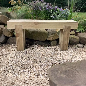May include: A light brown wooden bench constructed from thick, rectangular timber beams. The design features a long horizontal seat supported by two vertical legs joined with visible notches, showcasing a natural wood grain texture throughout.