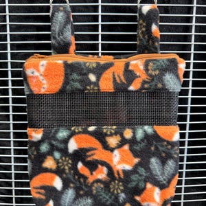 May include: A hanging pet feeder with a black mesh front and a zippered top. The feeder is decorated with a repeating pattern of orange foxes, white accents, and green foliage on a black background. Silver clips secure the feeder.
