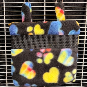 May include: A black fleece cage accessory with a heart pattern in yellow, blue, and pink. The cage accessory has a mesh front and is suspended by two metal clips.