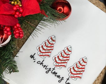 May include: White Christmas-themed kitchen towel with embroidered Christmas trees and the phrase "oh, christmas tree" in black script. The towel is decorated with red and green accents, and is surrounded by Christmas decorations.