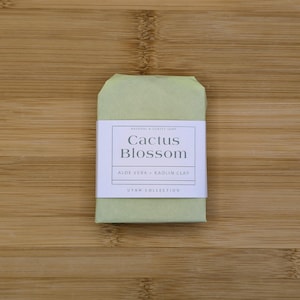 May include: A bar of natural soap wrapped in light green paper, with a white label that reads "Cactus Blossom." The label also includes "Aloe Vera + Kaolin Clay" and "Utah Collection."
