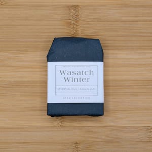 May include: A dark blue bar of natural soap wrapped in black paper. A white label reads "Wasatch Winter" with "Essential Oils + Kaolin Clay" and "Utah Collection" below. The soap is on a light brown wooden surface.