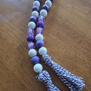 May include: A decorative beaded garland featuring alternating white and purple wooden beads, interspersed with textured purple beads. The garland is finished with two matching purple and white tassels.