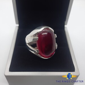May include: A silver ring featuring a large, oval-shaped, red gemstone set in the centre. The ring has a distinctive, intricate design with a silver band that wraps around the gemstone.