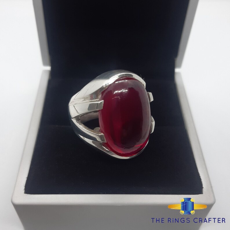 May include: A silver ring featuring a large, oval-shaped, red gemstone set in the centre. The ring has a distinctive, intricate design with a silver band that wraps around the gemstone.
