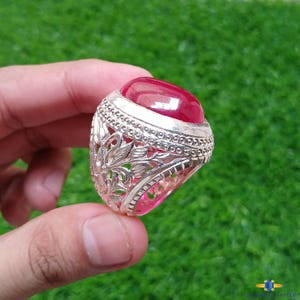 May include: A silver ring with an oval, deep red gemstone. The ring has an ornate, openwork design featuring floral and leaf patterns. The gemstone is set in a bezel setting, and the ring is held up by a hand against a green background.