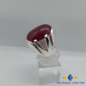 May include: A silver ring featuring a large, oval-shaped, red gemstone set in a silver setting. The ring is displayed on a white stand.