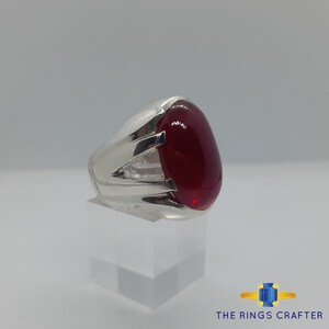 May include: A silver ring featuring a large, oval-shaped, red gemstone. The ring has a detailed design with three raised lines on the band.