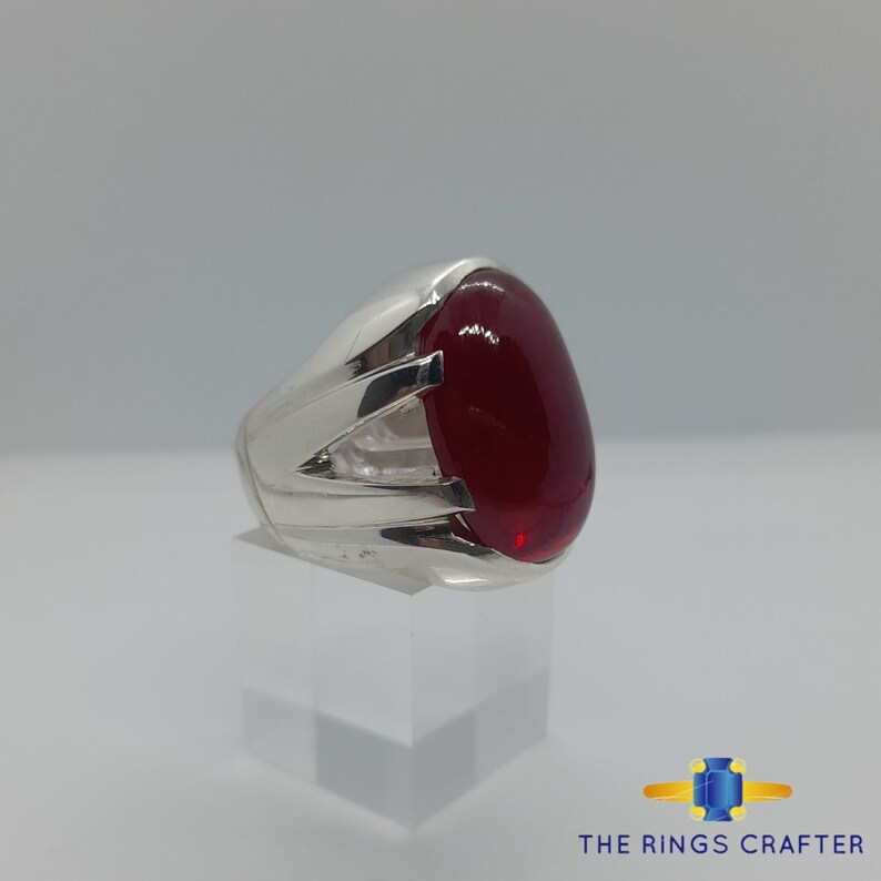 May include: A silver ring featuring a large, oval-shaped, red gemstone. The ring has a detailed design with three raised lines on the band.
