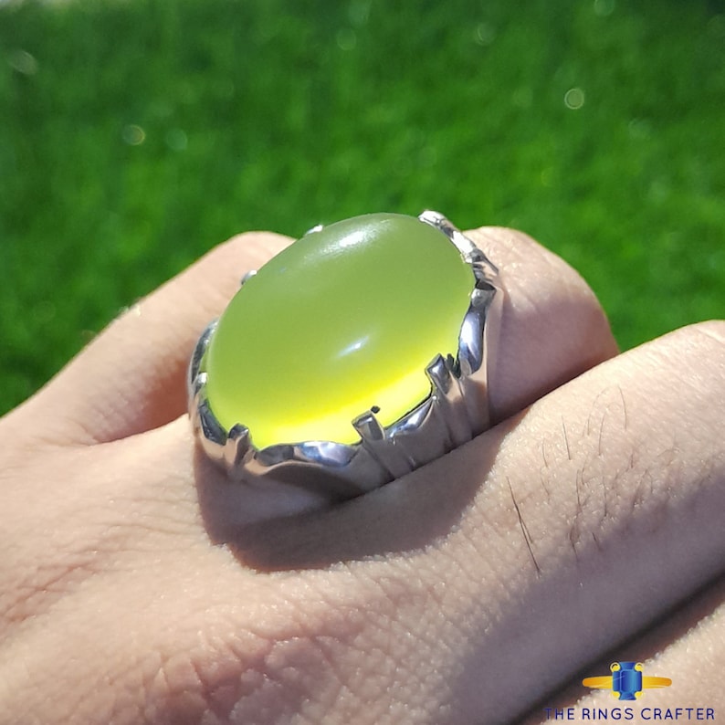 May include: A silver ring with a large, oval-shaped, light green gemstone. The ring is set in a silver band with a decorative design.