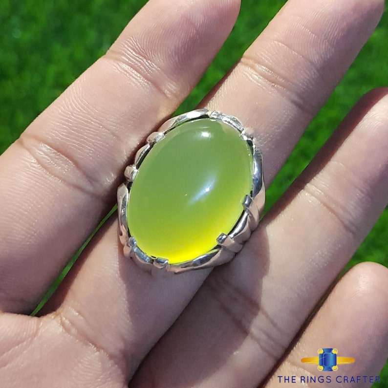 May include: A silver ring with a large, oval-shaped, light green gemstone. The ring has a detailed, intricate design around the stone.