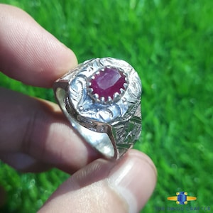 May include: A silver ring with an oval-shaped red gemstone set in a decorative silver setting. The ring has intricate floral designs engraved on the band.