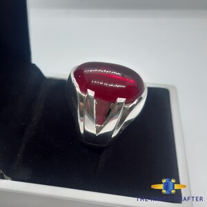 May include: A silver ring featuring a large, oval-shaped, red gemstone set in the centre. The ring has a V-shaped design on the band.