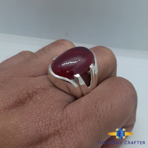 May include: A silver ring featuring a large, oval-shaped, red gemstone. The ring has a simple, modern design with a thick band.
