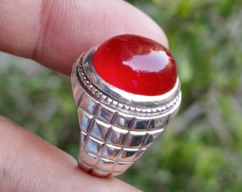 May include: A silver ring with a large, oval, red gemstone. The ring has a detailed, geometric design on the band and a textured bezel around the stone. The gemstone is polished and reflects light.