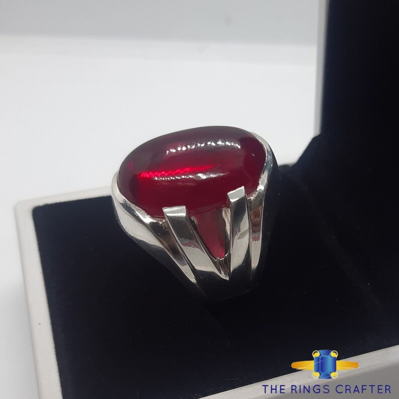 May include: A silver ring featuring a large, oval-shaped, deep red gemstone. The ring has a V-shaped design on the band.