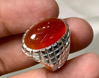 May include: A silver ring with a large, oval, reddish-orange gemstone. The ring's band features a textured, grid-like design. The gemstone has a polished, glossy surface. The ring is held between fingers, showcasing the jewelry.