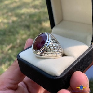 May include: A silver ring with a large, oval-cut, deep red gemstone. The ring has a textured, diamond-patterned band and is presented in a black jewelry box with a white interior. The ring is a statement piece.