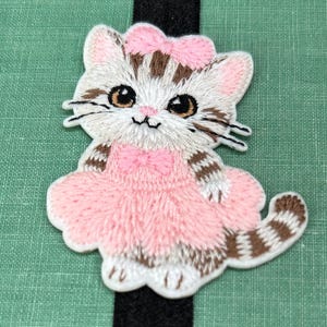 May include: Embroidered patch of a cute kitten wearing a pink dress and bow. The cat has brown and white stripes, with large brown eyes and whiskers. The patch is on a green background with a black stripe.