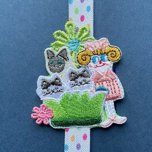 May include: A colorful bookmark with a green pot, three cats, and a woman with glasses and a pink dress. The bookmark is decorated with white polka dots and colorful dots.