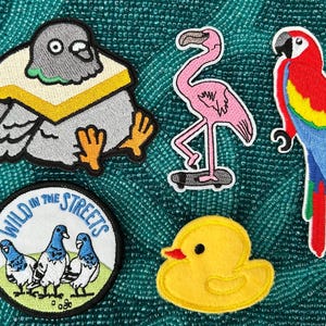 May include: A collection of embroidered patches featuring various birds and animals. Includes a pigeon with a sandwich, a pink flamingo on a skateboard, a colorful parrot, a yellow duck, and a patch with pigeons and the text "Wild in the Streets."