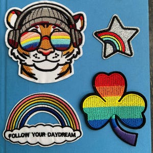 May include: A collection of embroidered patches on a blue background. Patches include a rainbow with the text "storms don't last forever", a tiger wearing sunglasses, a goose with a rainbow flag, a rainbow star, a rainbow clover, and a rainbow with the text "FOLLOW YOUR DAYDREAM".