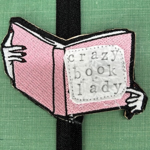 May include: A pink embroidered patch in the shape of an open book, with the words "crazy book lady" on a white label. The patch is held by white hands with black outlines. The background is green.