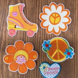 May include: Embroidered patches featuring a roller skate, two flowers, a peace sign, and a heart with the words "Flower Power." The patches are in shades of orange, yellow, pink, and blue, with white and yellow accents.
